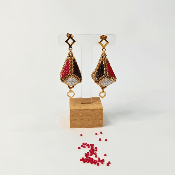 Orecchini pendenti forma a lanterna tridimensionale piena colore oro, nero, bianco e rosso su uno sfondo chiaro con perline in vetro rosse