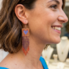 Orecchini di perline ramate iridescenti a frangia con cuore blu, indossato da donna sorridente con abito rosso, come sfondo la città