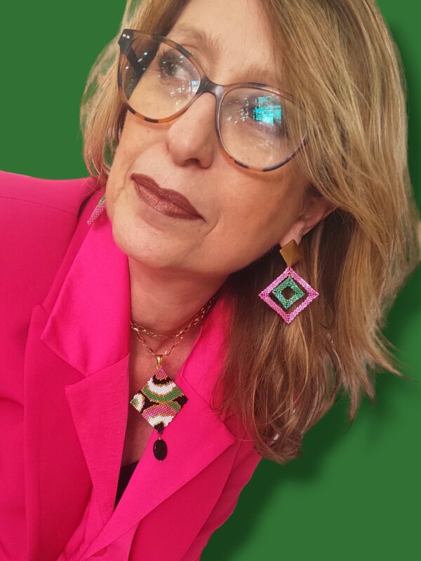 foto di donna bionda con occhiali che indossa un paio di orecchini pendenti con perno quadrato dorato. Il pendente è formato da due quadrati uno dentro l'altro. Verde il quadrato piccolo e rosa il quadrato grande. Indossa anache una collana corta con pendente quadrato e design anni 70 rosa, verde, bianco e nero. Indossa una giacca fucsia.