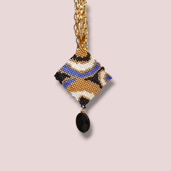 Sullo sfondo beige una collana con catena in acciaio dorato e ciondolo dal design anni 60/70 colore nero, bianco, oro, blu.