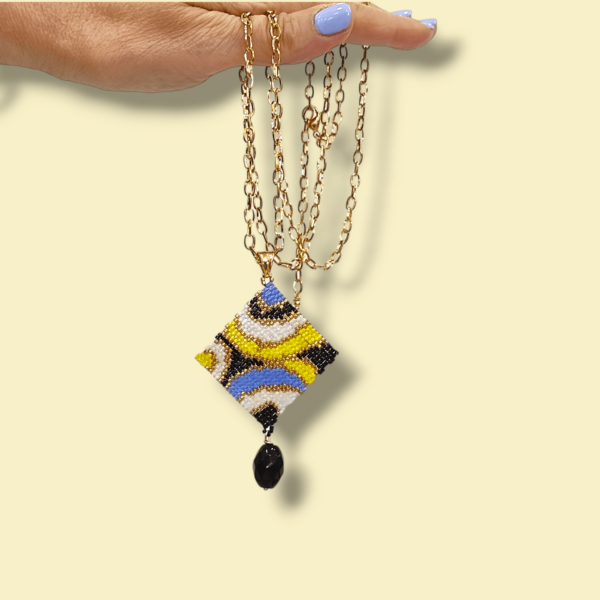Sullo sfondo giallino una collana con catena in acciaio dorato e ciondolo dal design anni 60/70 colore nero, bianco, oro, pervinca e giallo
