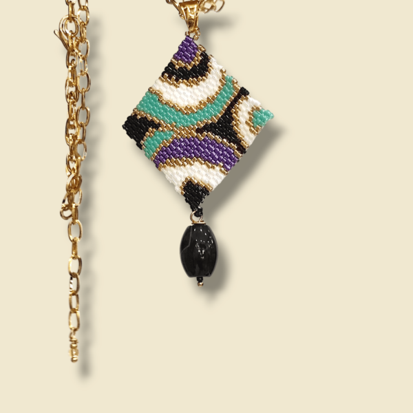 Sullo sfondo beige una collana con catena in acciaio dorato e ciondolo dal design anni 60/70 colore nero, bianco, oro, turchese e viola