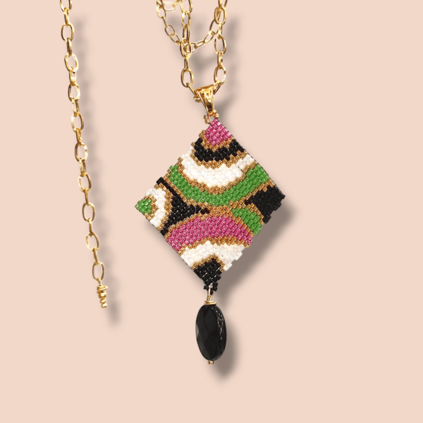 Sullo sfondo beige una collana con catena in acciaio dorato e ciondolo dal design anni 60/70 colore nero, bianco, oro, rosa e verde