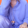 Su uno sfondo arancione dettaglio di donna piegata che indossa una camicia e pantaloni lilla, una collana con ciondolo quadrato.