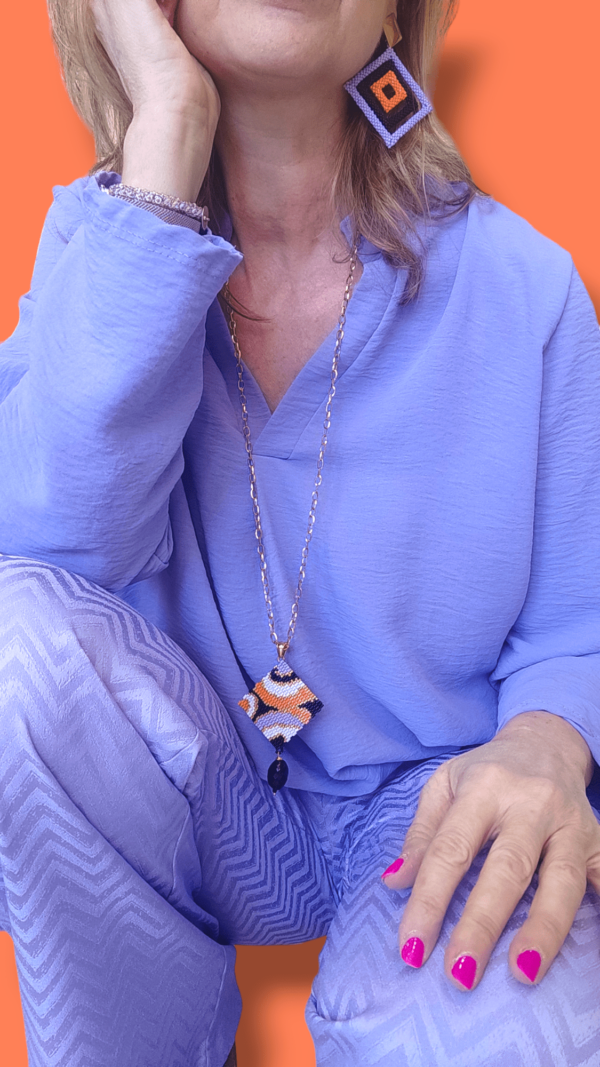 Su uno sfondo arancione dettaglio di donna piegata che indossa una camicia e pantaloni lilla, una collana con ciondolo quadrato.