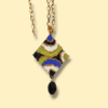 Sullo sfondo giallo tenue una collana con catena in acciaio dorato e ciondolo dal design anni 60/70 colore nero, bianco, oro, blu e verde lime