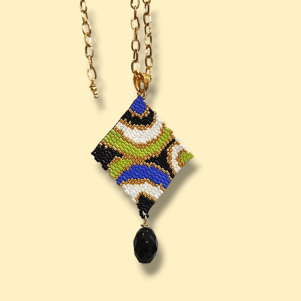 Sullo sfondo giallo tenue una collana con catena in acciaio dorato e ciondolo dal design anni 60/70 colore nero, bianco, oro, blu e verde lime