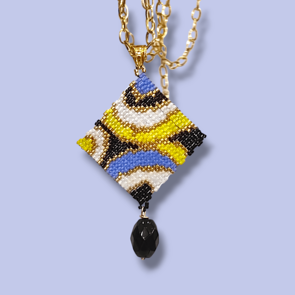 Sullo sfondo azzurro una collana con catena in acciaio dorato e ciondolo dal design anni 60/70 colore nero, bianco, oro, pervinca e giallo