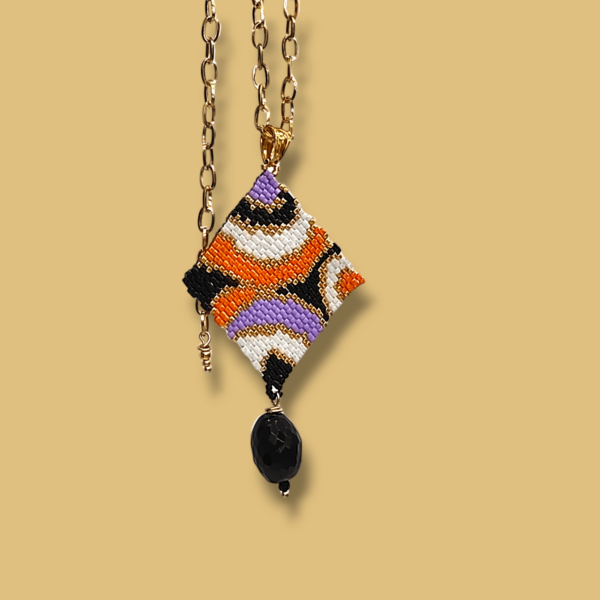 Sullo sfondo dorato una collana con catena in acciaio dorato e ciondolo dal design anni 60/70 colore nero, bianco, oro, lilla e arancione