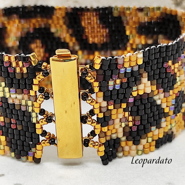 Inquadratura ravvicinata di un bracciale artigianale a fascia larga con motivo maculato leopardato, realizzato con perline Miyuki nere, oro e bronzo metallizzato. Al centro spicca la chiusura a baionetta in metallo dorato lucido.