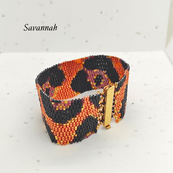Bracciale a fascia larga in perline Miyuki modello Savannah con motivo maculato arancione vibrante, corallo e nero opaco. In primo piano la chiusura rettangolare metallica in oro lucido su sfondo chiaro.