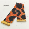 Bracciale a fascia larga in perline Miyuki modello Savannah con motivo maculato arancione vibrante, corallo e nero opaco. In primo piano la chiusura rettangolare metallica in oro lucido su sfondo chiaro.