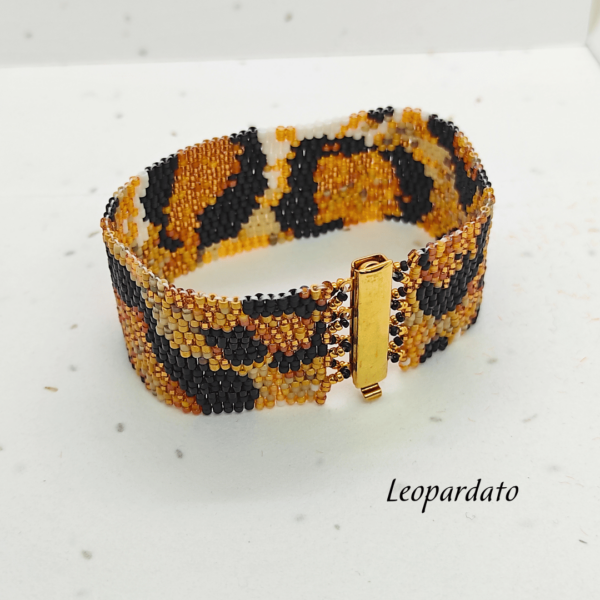 Bracciale rigido a fascia larga realizzato a mano con perline di vetro Miyuki con motivo leopardato animalier nei toni del nero, oro e marrone, dotato di chiusura a incastro in metallo dorato lucido su sfondo chiaro.