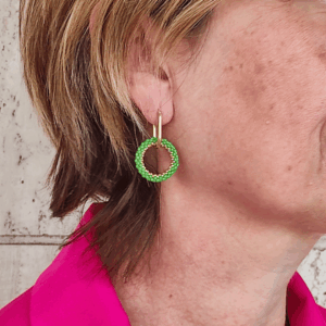Orecchini a cerchio con perno ovale in accaio dorato e cerchio di perline verde, indossato da donna con giacca fucsia