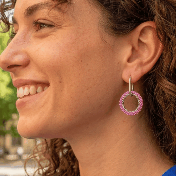 Orecchini a cerchio con perno ovale in accaio e cerchio di perline rosa, indossato da donna con maglia blu