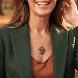Collana con ciondolo a forma di ghianda dai riflessi metallici rossi, blu e rame, realizzata a mano con micro perline di vetro e abbinata a una catenina dorata, indossata da donna con giacca verde