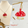 Collana fatta a mano con ciondolo a forma di funghetto rosso a pois bianchi, creata con un mosaico di perline di vetro e mostrata sia indossata su espositore sia in primo piano.
