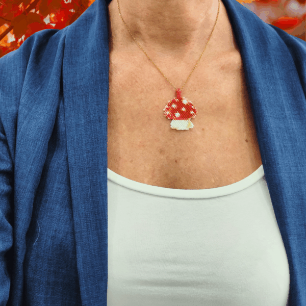 Collana fatta a mano con ciondolo a forma di funghetto rosso a pois bianchi, creata con un mosaico di perline di vetro e mostrata sia indossata su espositore sia in primo piano. Indossato