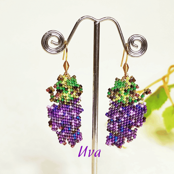 Orecchini fatti a mano a forma di grappoli d'uva viola con foglioline verdi, realizzati con un mosaico di perline di vetro e appesi a un espositore metallico.