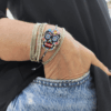 Inquadratura ravvicinata del bracciale teschio messicano Calaveras indossato al polso, parzialmente inserito nella tasca di un paio di jeans chiari. Il pendente centrale mostra una tessitura geometrica fitta di micro-perline multicolor con dettagli fucsia, azzurri e gialli, rifinito con nodi in macramè grigio scuro. Abbinato a bracciali rigidi multifilo, incarna perfettamente lo spirito libero, dinamico ed eclettico di Vanitage.
