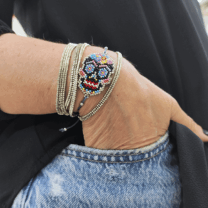 Inquadratura ravvicinata del bracciale teschio messicano Calaveras indossato al polso, parzialmente inserito nella tasca di un paio di jeans chiari. Il pendente centrale mostra una tessitura geometrica fitta di micro-perline multicolor con dettagli fucsia, azzurri e gialli, rifinito con nodi in macramè grigio scuro. Abbinato a bracciali rigidi multifilo, incarna perfettamente lo spirito libero, dinamico ed eclettico di Vanitage.