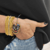 Inquadratura ravvicinata del bracciale teschio messicano Calaveras indossato al polso, parzialmente inserito nella tasca di un paio di jeans chiari. Il pendente centrale mostra una tessitura geometrica fitta di micro-perline multicolor con dettagli fucsia, azzurri e gialli, rifinito con nodi in macramè grigio scuro. Abbinato a bracciali rigidi multifilo, incarna perfettamente lo spirito libero, dinamico ed eclettico di Vanitage.