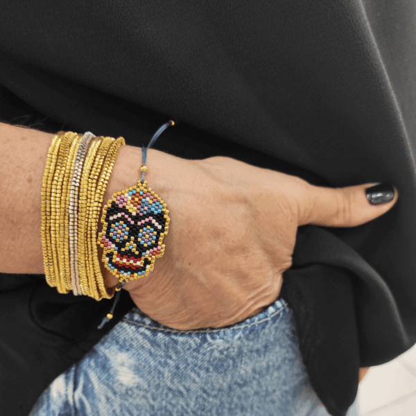 Inquadratura ravvicinata del bracciale teschio messicano Calaveras indossato al polso, parzialmente inserito nella tasca di un paio di jeans chiari. Il pendente centrale mostra una tessitura geometrica fitta di micro-perline multicolor con dettagli fucsia, azzurri e gialli, rifinito con nodi in macramè grigio scuro. Abbinato a bracciali rigidi multifilo, incarna perfettamente lo spirito libero, dinamico ed eclettico di Vanitage.