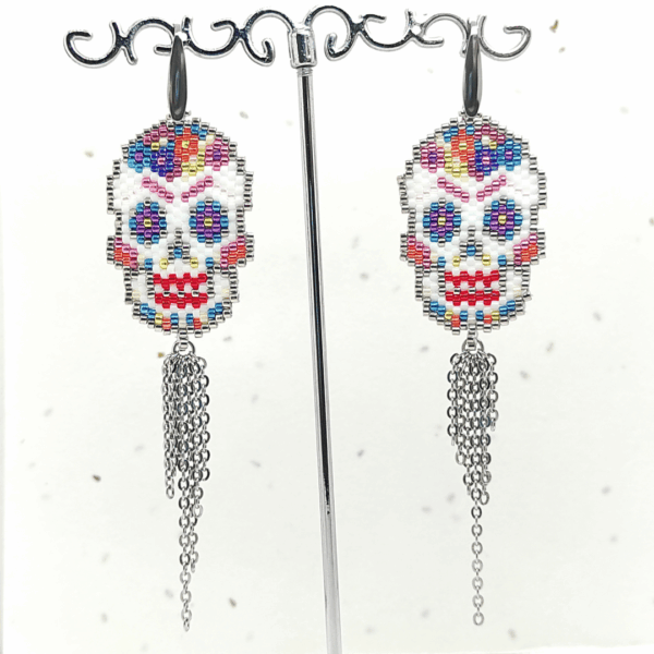 Orecchini pendenti Vanitage con teschio messicano in micro-perline multicolor e lunghe frange di catenelle argentate a movimento fluido, indossati al lobo.