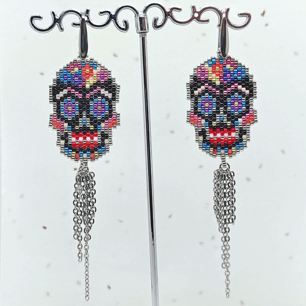 Orecchini pendenti Vanitage con teschio messicano in micro-perline multicolor e lunghe frange di catenelle argentate a movimento fluido, indossati al lobo.